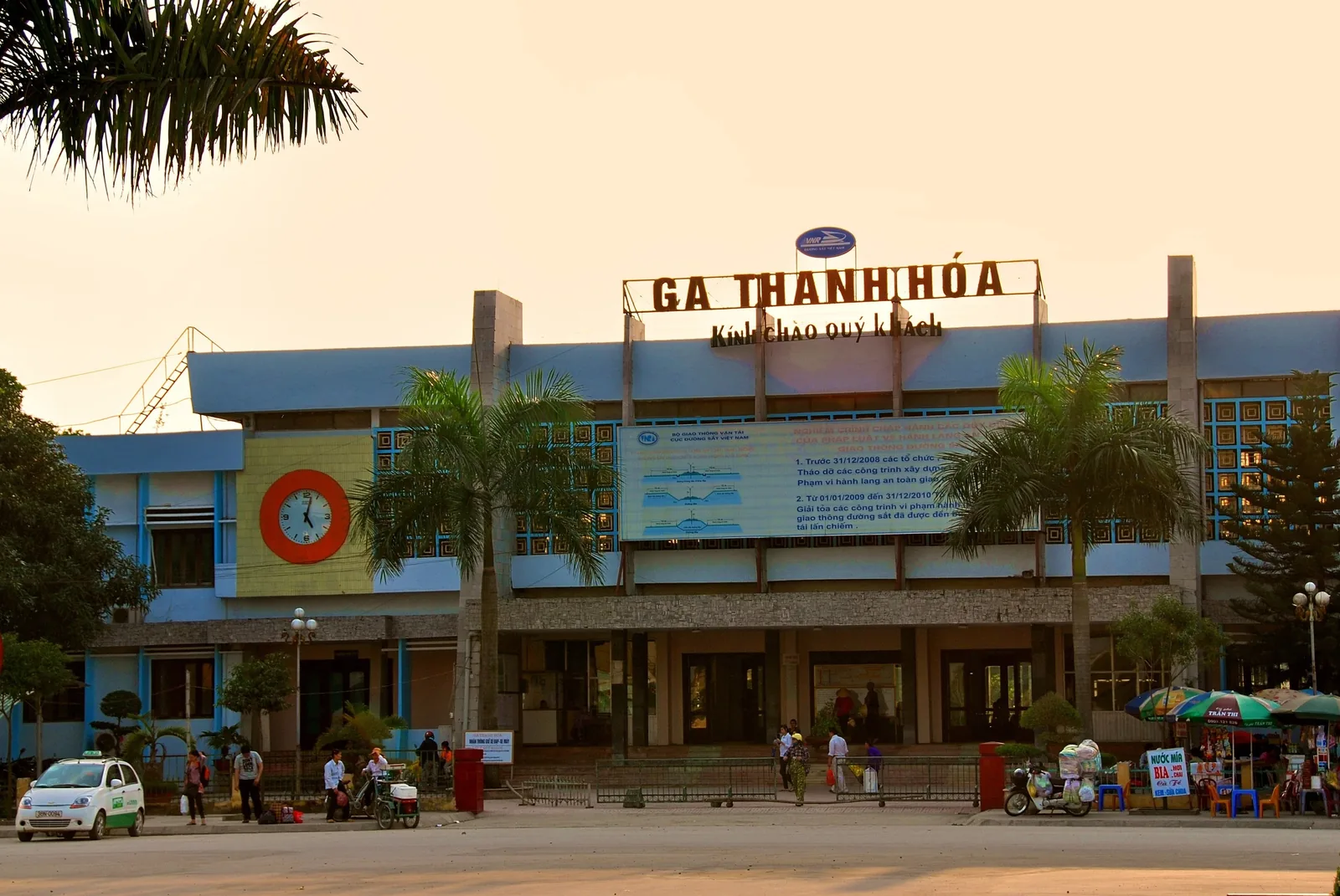 Mặt tiền ga Thanh Hóa ở Việt Nam lúc chiều tối, với hàng cọ và người bán hàng bên ngoài.