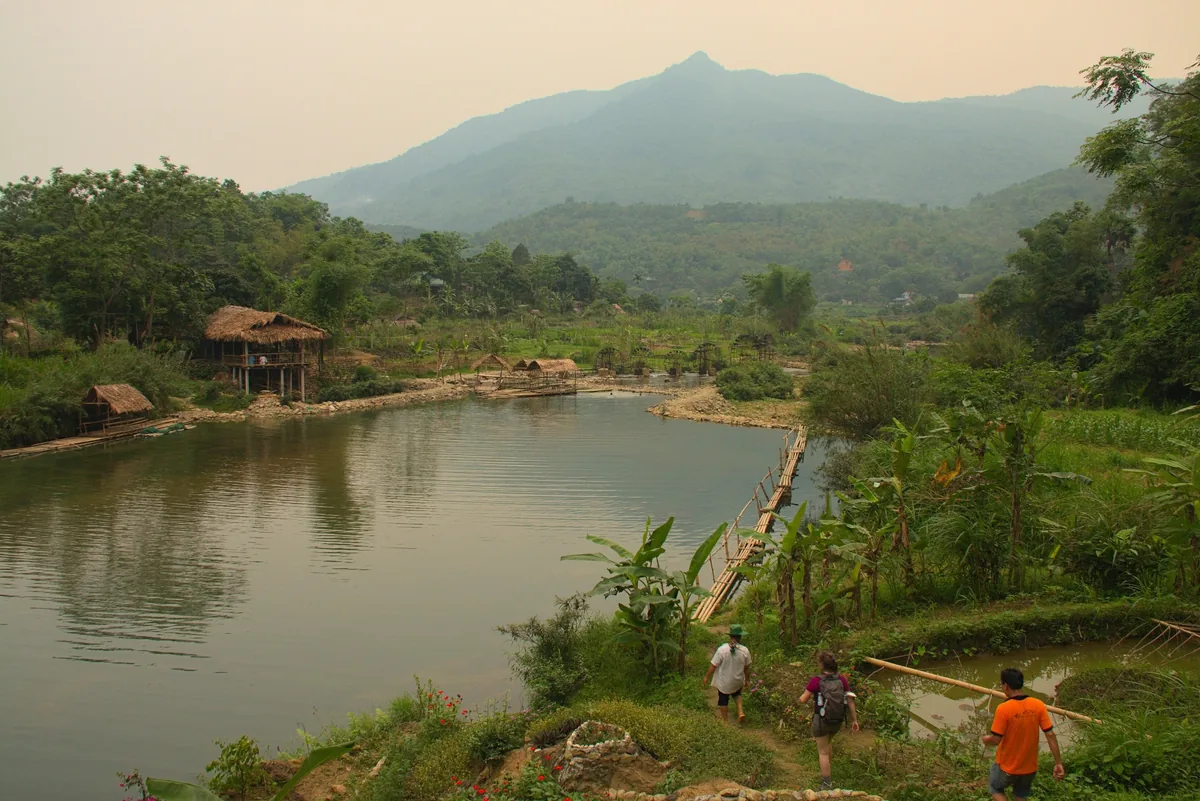 Người đi bộ dọc con đường bên sông gần những căn nhà tre và núi ở Pù Luông, Việt Nam.