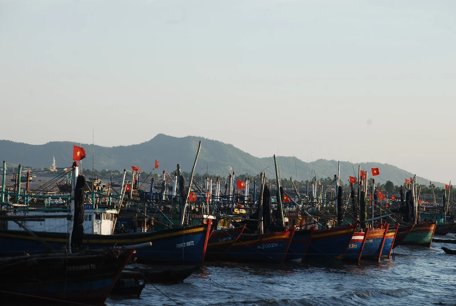 Nhiều thuyền cá Việt Nam cắm cờ đỏ đậu san sát tại cảng ven biển dưới chân đồi núi mờ xa.